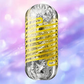 A transparent Tenga Spinner DX 02 Bumps male masturbator by Tenga, featuring metallic crumpled material and a yellow spiral, is set against a blurred pastel background in pink, purple, and blue tones.
